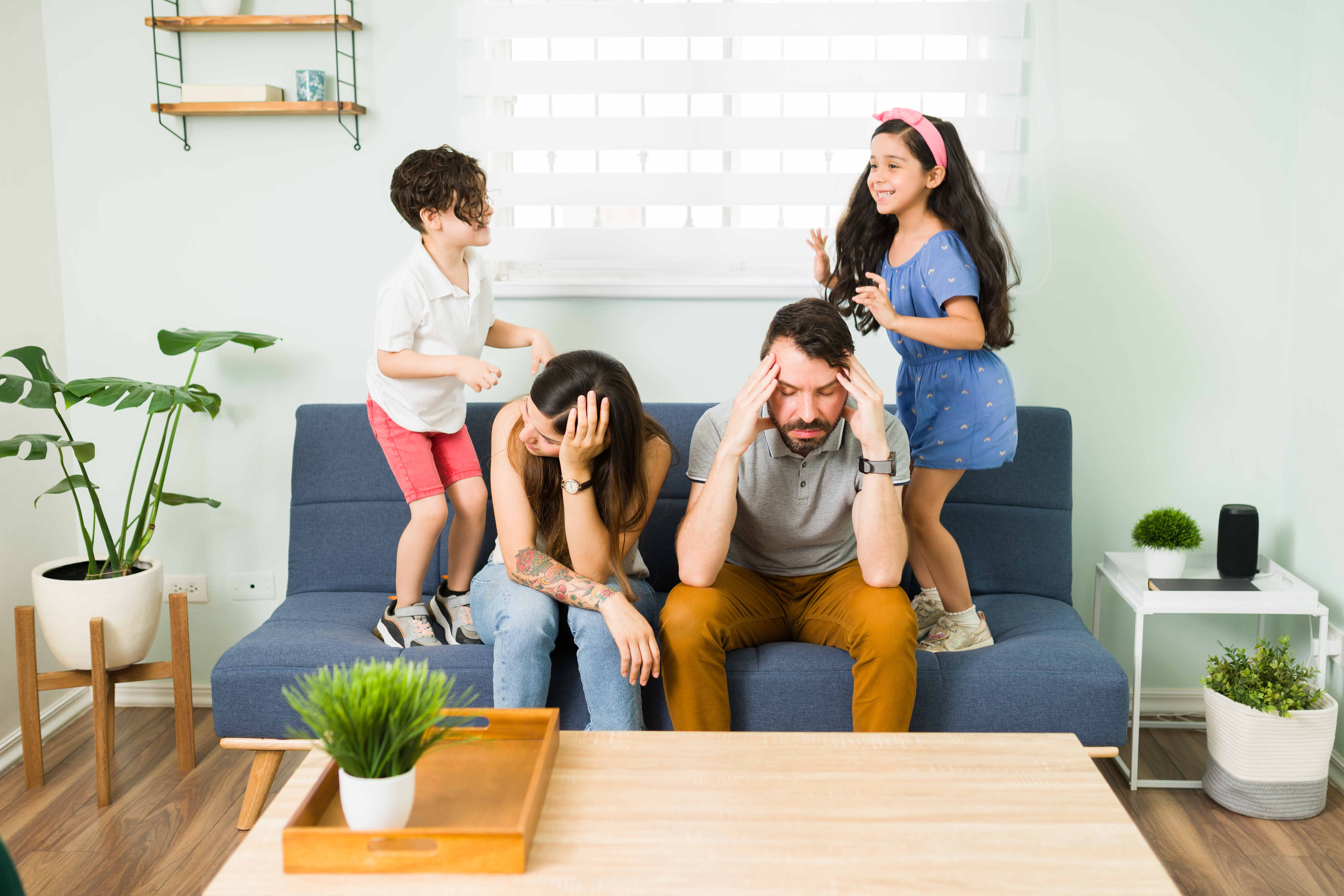 Stressed parents on a couch while their two children jump around them