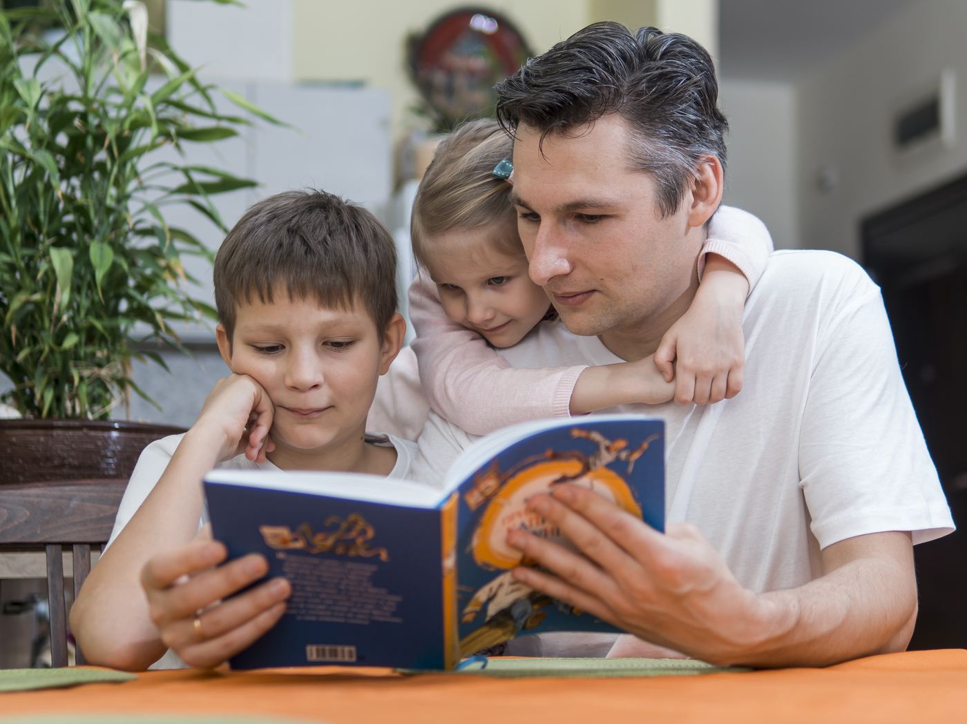 A father reading a book with his two young children at the kitchen table