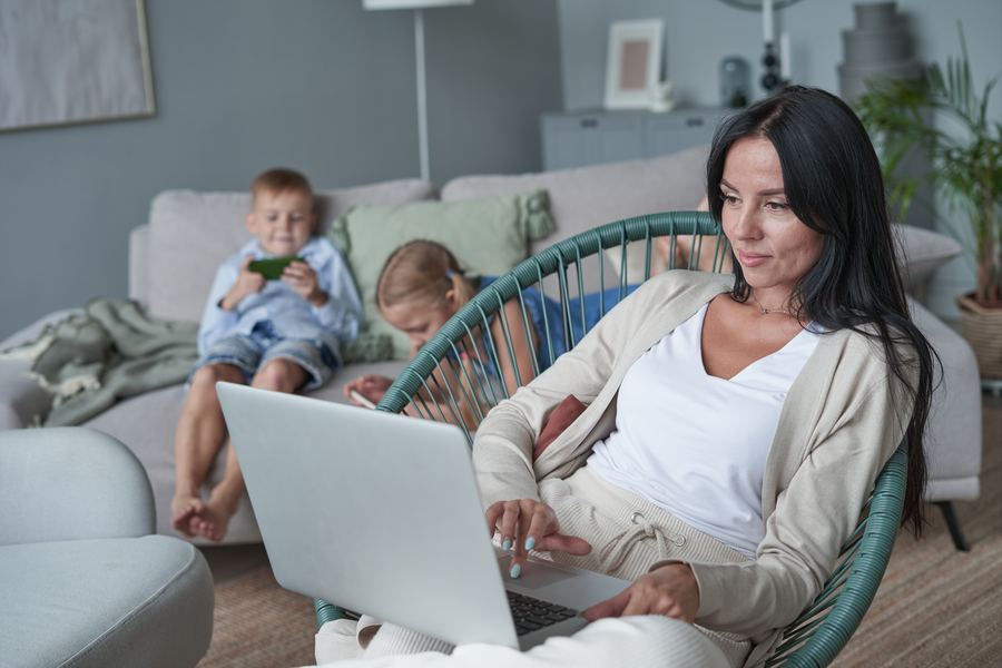 A mother using a laptop while her children relax on the sofa